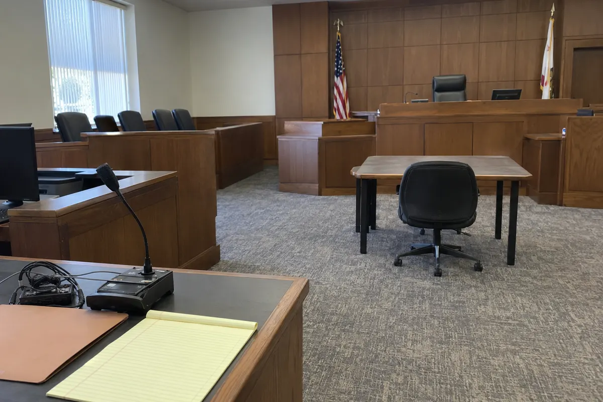 An empty courtroom in flat daylight seen from the corner of a clerk's desk: a small black audio recorder with a gooseneck microphone and a tangle of cable sits beside a closed manila folder and a blank yellow legal pad. Across the carpet, an empty office chair stands at a bare table in front of an empty judge's bench. No people are in the frame.