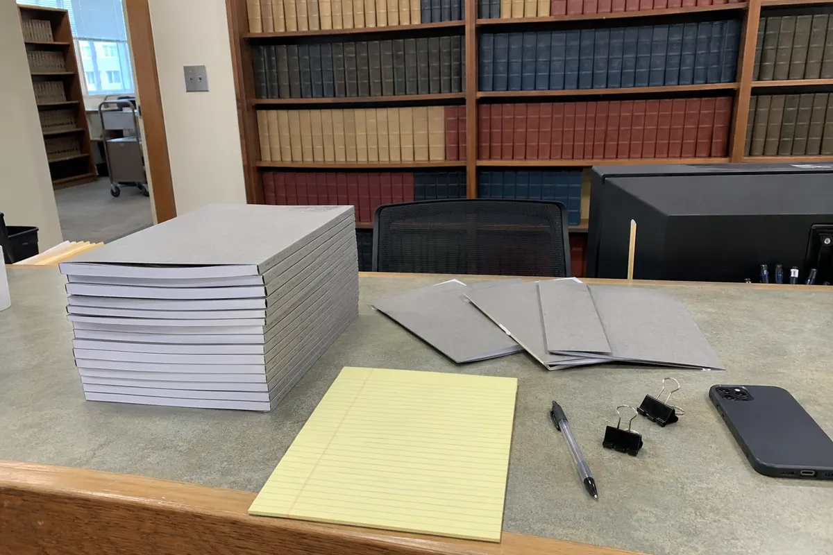 A clerk's filing counter in flat daylight: a tall stack of identical bound briefs with plain gray covers, a few more fanned out beside it, a blank yellow legal pad, a pen, two binder clips and a phone lying face down, with shelves of law reports behind. No people are in the frame.