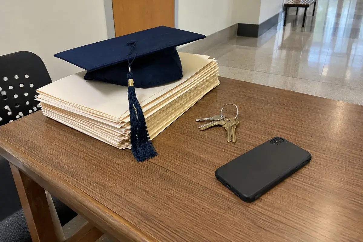 A navy graduation cap with its tassel rests on a thick stack of unlabeled manila folders on a wooden table in a bright school hallway, beside a ring of keys and a phone lying face down. No people are in the frame.