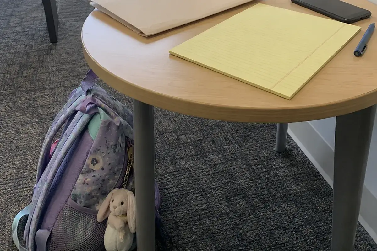 A round wooden table in a carpeted waiting area in flat daylight: a closed manila folder, a blank yellow legal pad, a pen and a phone lying face down on the tabletop, and a child's lavender backpack with a small stuffed rabbit clipped to it leaning against a table leg. No people are in the frame.