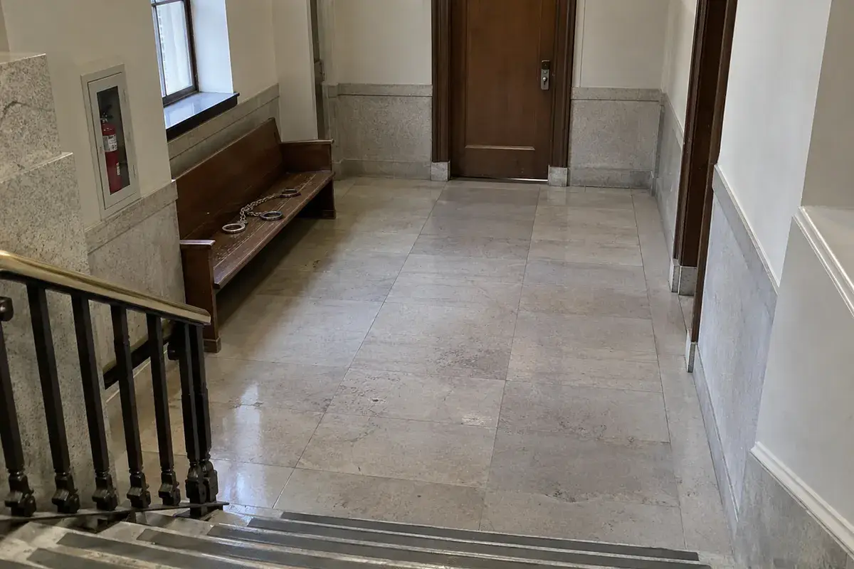 An empty courthouse hallway in flat daylight seen from the top of a stone staircase: a set of steel handcuffs joined by a chain lies on a wooden bench against the wall, beside a fire extinguisher cabinet, with a closed wooden door at the end of the corridor. No people are in the frame.
