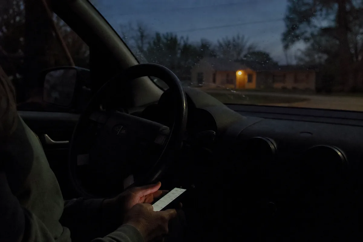 Las manos de una mujer sobre su regazo sosteniendo un teléfono, vistas desde el asiento del copiloto de un auto estacionado al atardecer.