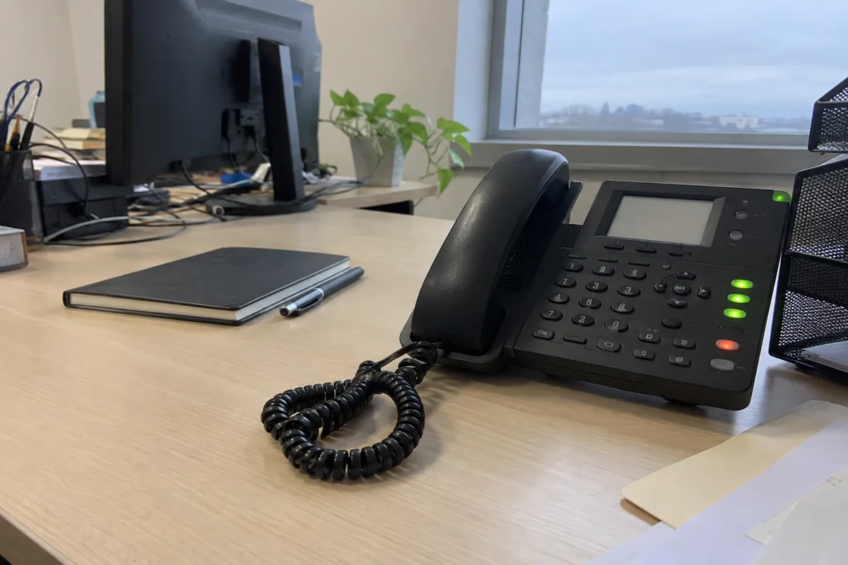 A multi-line office desk phone on a light wood desk, photographed off-center in flat overcast daylight from a window, with the corded handset resting in its cradle, a coiled cord looped on the desk, several small indicator lights along the phone's base, a closed plain notepad and a pen beside it, and a blurred computer monitor and a small potted plant in the background. No people are in the frame.