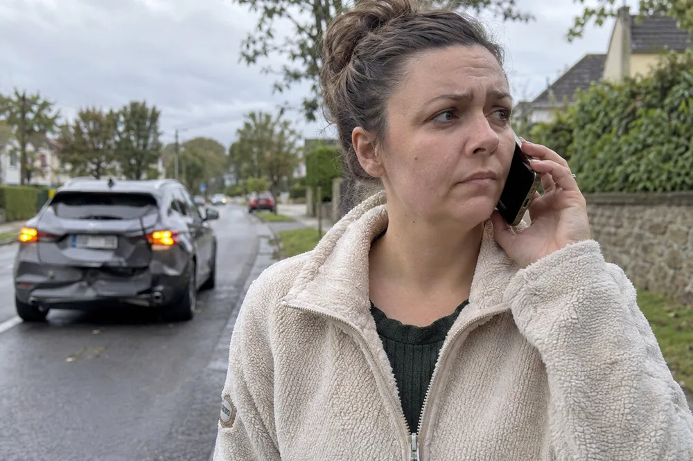A hand holding a phone on a roadside call, with a car with a dented bumper behind.